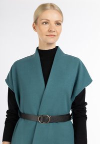 Teal wikkelvest van zacht materiaal, met korte mouwen en een zwarte leren riem met een ronde gouden gesp, gelaagd over een zwarte top.