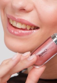 Gros plan d'une personne souriante tenant un gloss à lèvres KIKO Milano rose près de ses lèvres brillantes.