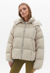Beige gewatteerde jas met een ritssluiting aan de voorkant, hoge kraag en grote gestepte secties. Gemaakt van gladde stof met een zachte glans en elastische manchetten.