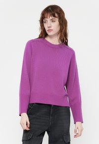 Pull en tricot violet avec un col rond, des poignets et un ourlet côtelés. Coupe ample avec des manches longues et une texture lisse. Pantalon sombre porté en dessous.