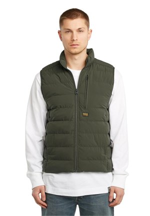 Green puffer vest with quilted design, high collar, full front zip, and logo patch. Worn over a white long-sleeve shirt. Casual fit.