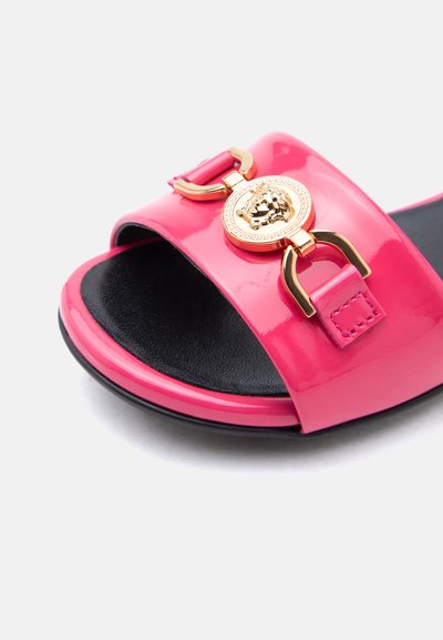 Sandale rose à bout ouvert avec semelle intérieure noire et emblème décoratif doré représentant une tête de lion sur la lanière supérieure.