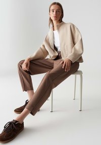Femme assise sur un tabouret blanc portant une veste zippée beige, une chemise blanche, un pantalon marron et des baskets marron foncé, sur fond gris clair.