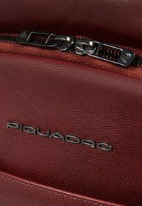 Sac à dos en cuir bordeaux à finition texturée, doté d'une fermeture éclair noire et d'un logo métallique "Piquadro" à l'avant.