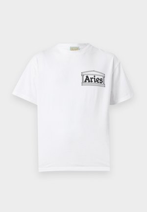 Bărbat cu păr creț purtând un tricou alb cu logo-ul "Berbec", blugi gri închis cu șnur și pantofi negri eleganți, stând în fața unui fundal alb.