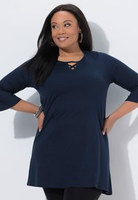Uśmiechnięta kobieta o długich ciemnych włosach, ubrana w granatową tunikę z rękawami do łokcia i geometrycznym dekoltem, z rękami opartymi na biodrach.