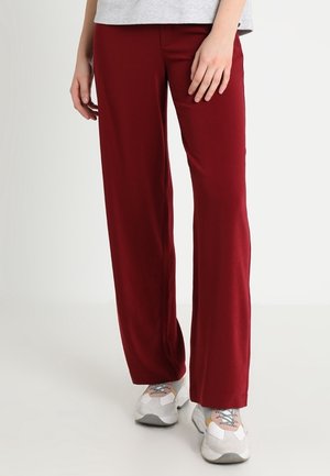 Personne portant un pantalon ample rouge foncé et des baskets blanches, debout sur un fond clair uni.