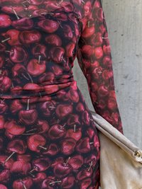 Langermet kjole med et rødt kirsebærmønster på en mørk bakgrunn, laget av elastisk stoff, med en drapert design og en glatt beige håndveske.