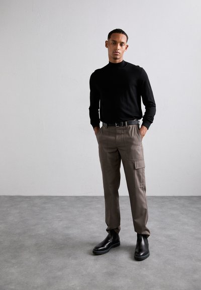 Schwarzes Langarm-Turtleneck, beige Cargo-Hosen mit Seitentaschen und schwarze Stiefeletten, vor einem minimalistischen Hintergrund.