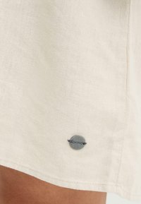 Tissu beige clair avec une petite étiquette métallique ronde cousue près de l'ourlet, affichant un nom de marque partiellement visible.