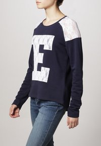 Maglione blu navy con maniche lunghe, dettagli in pizzo sulle spalle, una grande "E" bianca sul davanti e un taglio rilassato.