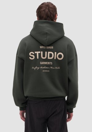 Hombre con sudadera con capucha verde oscuro con un gran texto beige que dice "STUDIO GARMENTS" y texto más pequeño en la parte trasera, combinado con pantalones de rayas finas.