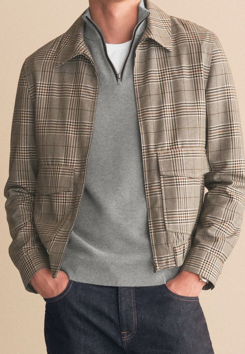 Karierte Jacke mit einem hellbraunen und beigen Muster aus Stoff. Sie hat einen klassischen Kragen und zwei Fronttaschen und wird über einem grauen Pullover getragen.