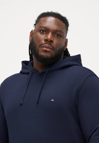 Granatowy sweatshirt z kapturem wykonany z miękkiego materiału, z małym logo w czerwonych, białych i niebieskich akcentach na piersi.