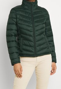 Vihreä pehmustettu takki, jossa on korkea kaulus, teksturoitu tikattu muotoilu, etuvetoketju ja sivutaskut. Yhdistetty vaaleanbeigeihin housuihin.