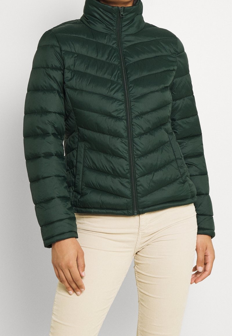 Vihreä pehmustettu takki, jossa on korkea kaulus, teksturoitu tikattu muotoilu, etuvetoketju ja sivutaskut. Yhdistetty vaaleanbeigeihin housuihin.