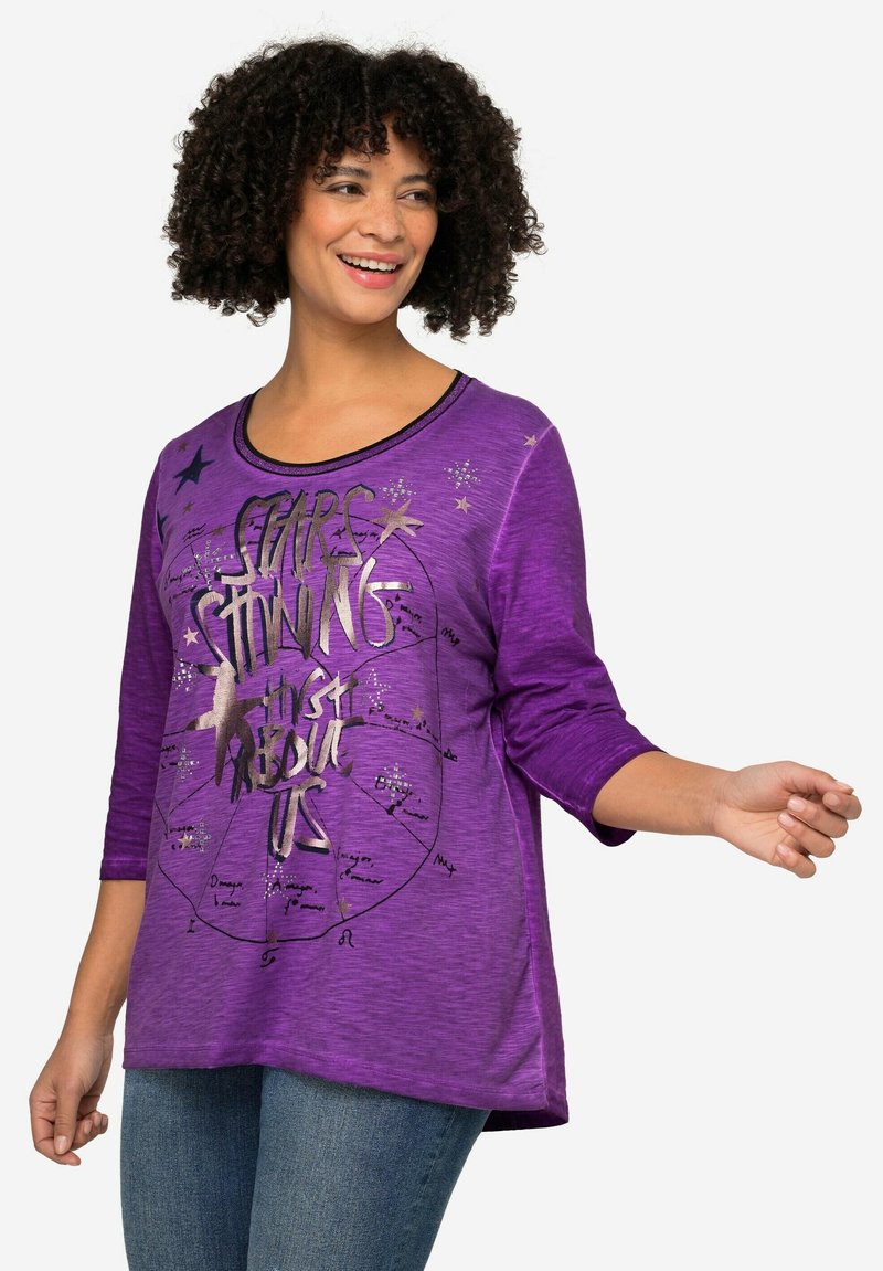 Camisa morada de manga tres cuartos con texto gráfico metálico y motivos de estrellas. Presenta un corte relajado y un escote redondeado.