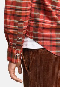 Camicia a quadri rossa a maniche lunghe con bottoni sui polsini, caratterizzata da varie tonalità di rosso, verde e marrone, indossata sopra pantaloni in velluto a coste marroni.