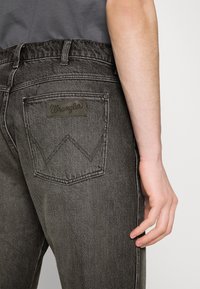 Jeans Wrangler grii cu un finisaj texturat. Prezintă un buzunar posterior cu un patch logo și un model de cusătură distinctiv în formă de 'W'.