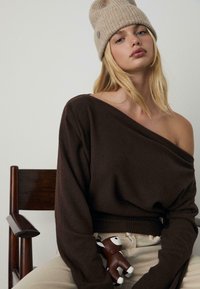 Brauner schulterfreier Pullover aus weichem Material, getragen mit cremefarbenen Hosen. Eine beigefarbene gerippte Mütze und ein kleines braunes Spielzeug sind zu sehen.