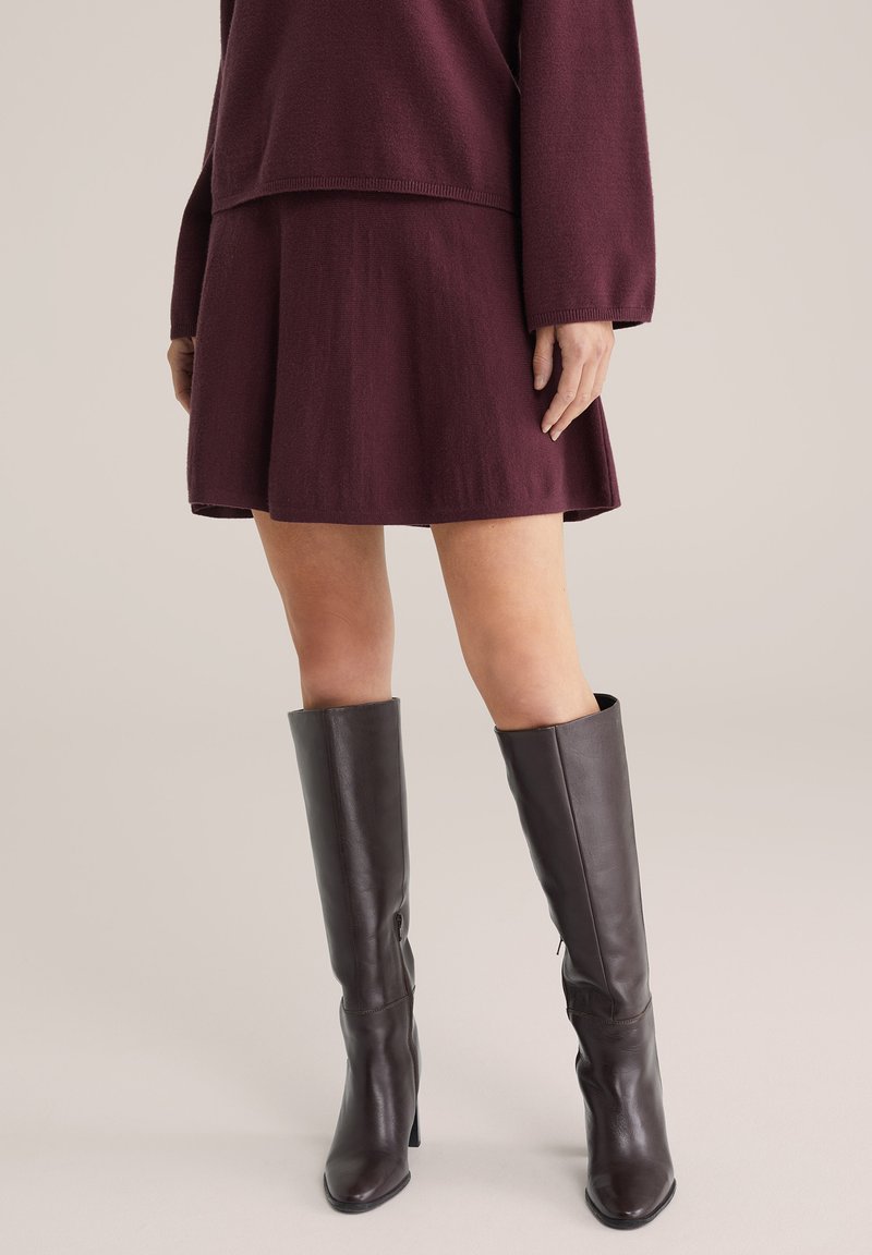 Burgunderfarbener Strickpulli und passender, ausgestellter Rock, kombiniert mit kniehohen braunen Lederstiefeln. Glatte Texturen mit minimaler Detailverarbeitung.