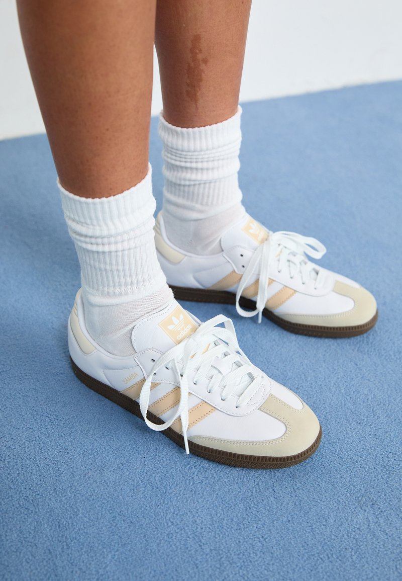 Fehér és bézs Adidas Samba tornacipők kerek orrakkal, fűzős kialakítással és texturált barna talppal, fehér bordás zoknikkal párosítva.