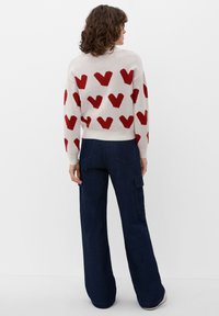 Maglione lavorato a maglia con motivi a cuore rossi su uno sfondo chiaro, abbinato a pantaloni in denim scuri a gamba larga con tasche laterali e una finitura testurizzata.