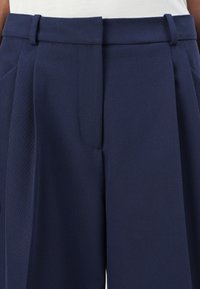 Mörkblå byxor med en texturerad tyg. Har flat front, veck, bältesöglor och dragkedja. Lämpliga för formell eller smart avslappnad klädsel.