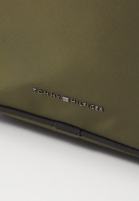 Oliven grønn stoffveske med en jevn tekstur, svarte detaljer og en sølvfarget "TOMMY HILFIGER"-logo preget på siden.