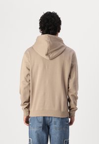 Hættetrøje i lys beige, lavet af blødt materiale. Har en afslappet pasform, ribbede manchetter og kant. Bæres med blå slidte denimjeans.