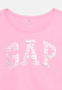 Otroška roza majica s srebrnimi črkami v obliki metulja, ki na sprednji strani tvorijo besedo "GAP", z okroglim izrezom, izdelano v Indiji.