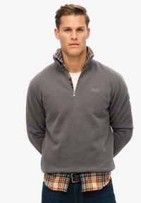 Sudadera de forro polar gris con cuello con cremallera, superpuesta a una camisa de cuadros en tonos rojo, amarillo y azul marino. Textura suave, diseño casual.