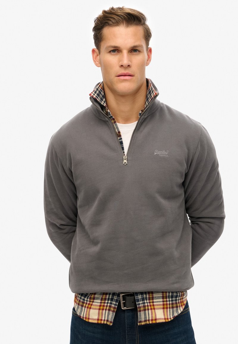 Sudadera de forro polar gris con cuello con cremallera, superpuesta a una camisa de cuadros en tonos rojo, amarillo y azul marino. Textura suave, diseño casual.