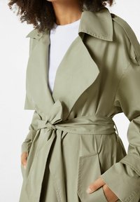 Kapsářový kabát v olivově zelené barvě, vyrobený z hladkého materiálu. Má límec, pás na zavázání a boční kapsy. Dlouhé rukávy s ohrnutými manžetami.