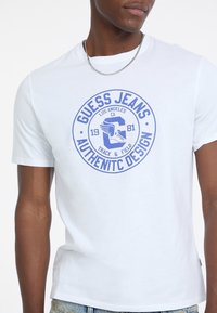 Mężczyzna w białej koszulce z niebieskim okrągłym logo "Guess Jeans Authentic Design" oraz grafiką skrzydlatej buty na klatce piersiowej.