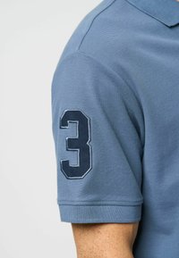 Zila polo krekls ar teksturētu audumu, īsām piedurknēm un tumši zilā izšūtā skaitļa "3" uz kreisās piedurknes.