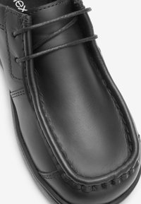 Chaussure noire en cuir à enfiler avec des coutures visibles, bout rond et détail lacé. Texture lisse et design minimaliste.