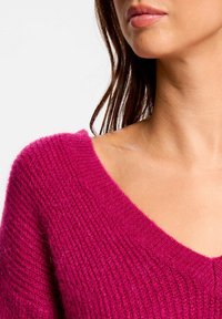 Frau in einem tief rosa gestrickten V-Ausschnitt-Pullover, der Schulter, Hals und Untergesicht zeigt, mit heller Haut und braunem Haar.