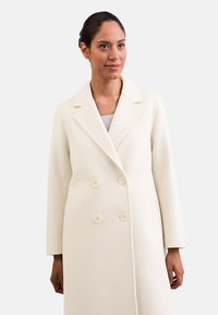 Cappotto doppiopetto in crema con rever a punta, maniche lunghe e due tasche frontali. Presenta bottoni tondi abbinati e una texture liscia.