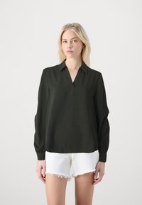 Blusa verde scuro con scollatura a V, maniche lunghe con dettagli ai polsini, realizzata in tessuto testurizzato, abbinata a pantaloni corti bianchi sfilacciati.
