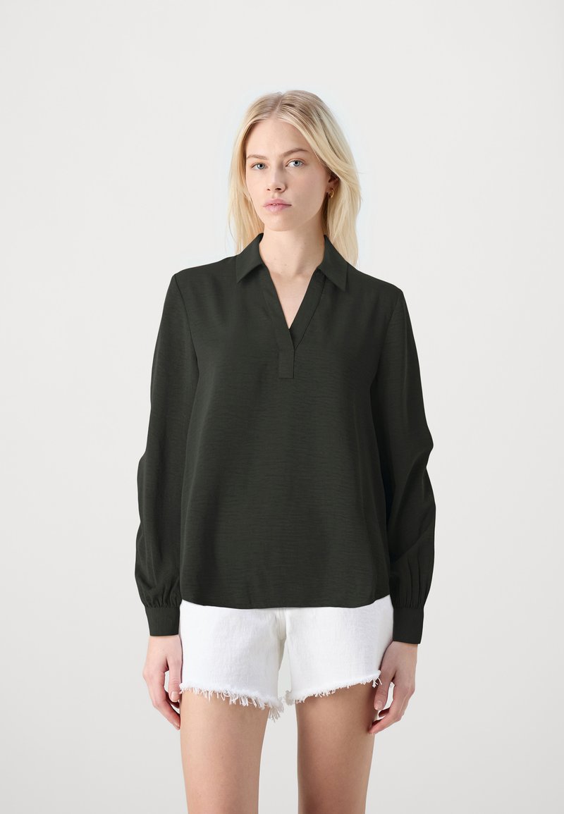 Blusa verde scuro con scollatura a V, maniche lunghe con dettagli ai polsini, realizzata in tessuto testurizzato, abbinata a pantaloni corti bianchi sfilacciati.