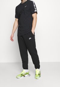 Nike svart mönstrad T-shirt med vitt logo, tillsammans med svarta sweatpants med ett logo. Klara gröna och gula träningsskor.