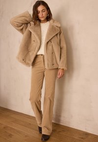 Veste beige en fausse fourrure à double boutonnage avec de gros boutons, associée à un pantalon en velours côtelé beige clair et un pull texturé crème. Bottes marron.