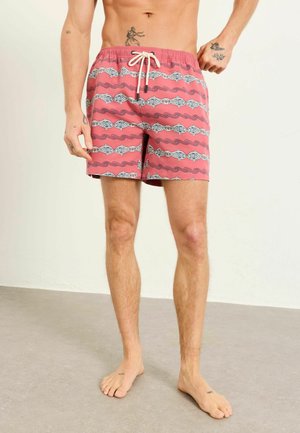 Muž v červených plavkách s bielymi a čiernymi rybími vzormi, stojaci naboso na sivej podlahe pred obyčajnou stenou.