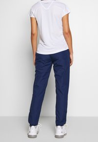 Bílý top s krátkým rukávem a tmavě modré kalhoty z lehkého materiálu, které mají volný střih. Celkový dojem doplňují bílé sportovní boty.