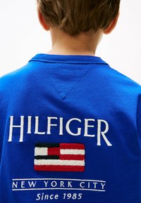 Blauer Sweatshirt mit weißem gesticktem "HILFIGER"-Text und einem strukturierten Flaggenmotiv in Rot, Weiß und Schwarz, mit "NEW YORK CITY" darunter.