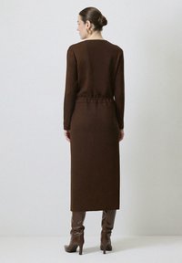 Robe longue en tricot marron avec une silhouette droite, un col rond et un détail de taille froncé, associée à des bottes marron jusqu'aux genoux.