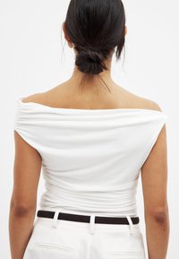 White off-shoulder top with gathered fabric detail at the back, smooth texture, paired with high-waisted trousers and a black belt.