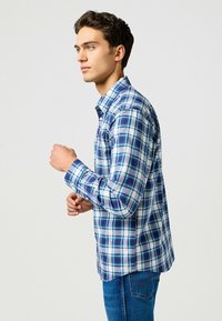 Camicia a quadri blu e bianchi con maniche lunghe e colletto a pulsante, realizzata in tessuto leggero. Indossata con jeans blu, vista di lato.