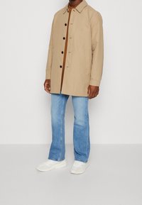 Beige buitenjack met een kraag, knoopsluiting aan de voorkant en twee zijzakken, gedragen over een bruine top en lichtblauwe jeans. Witte sneakers zijn te zien.